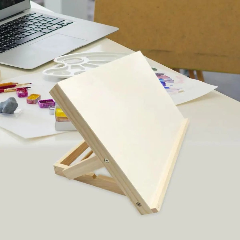Schuin gezette houten tafelezel op een witte tafel met aquarelverf, potloden en een laptop erachter