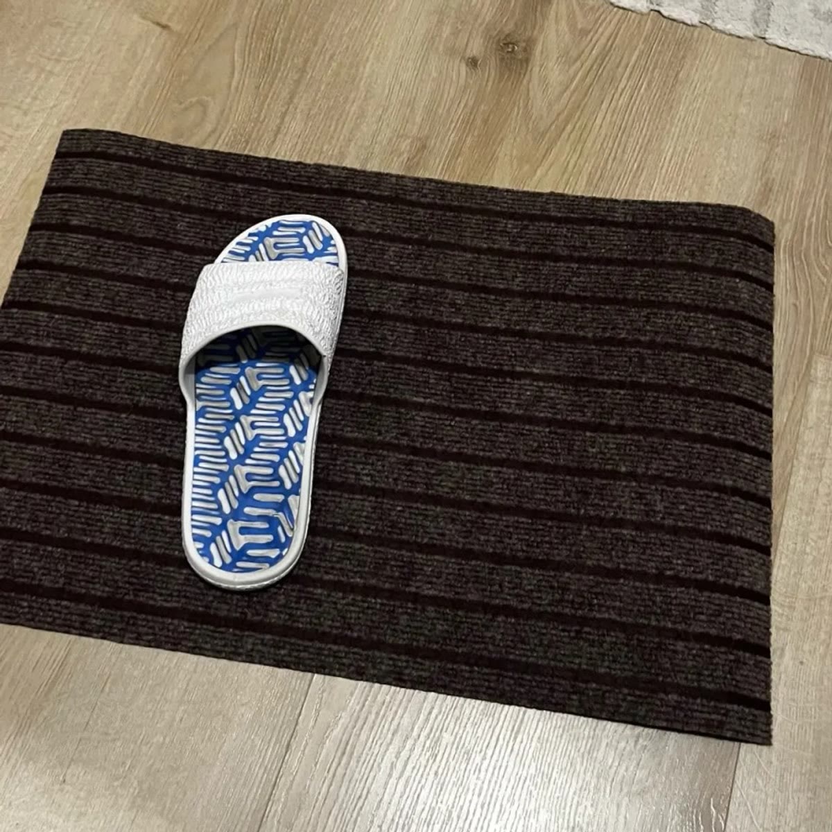 De grijsbruine deurmat op een lichte vloer, met een wit-blauwe slipper erop die de grootte laat zien