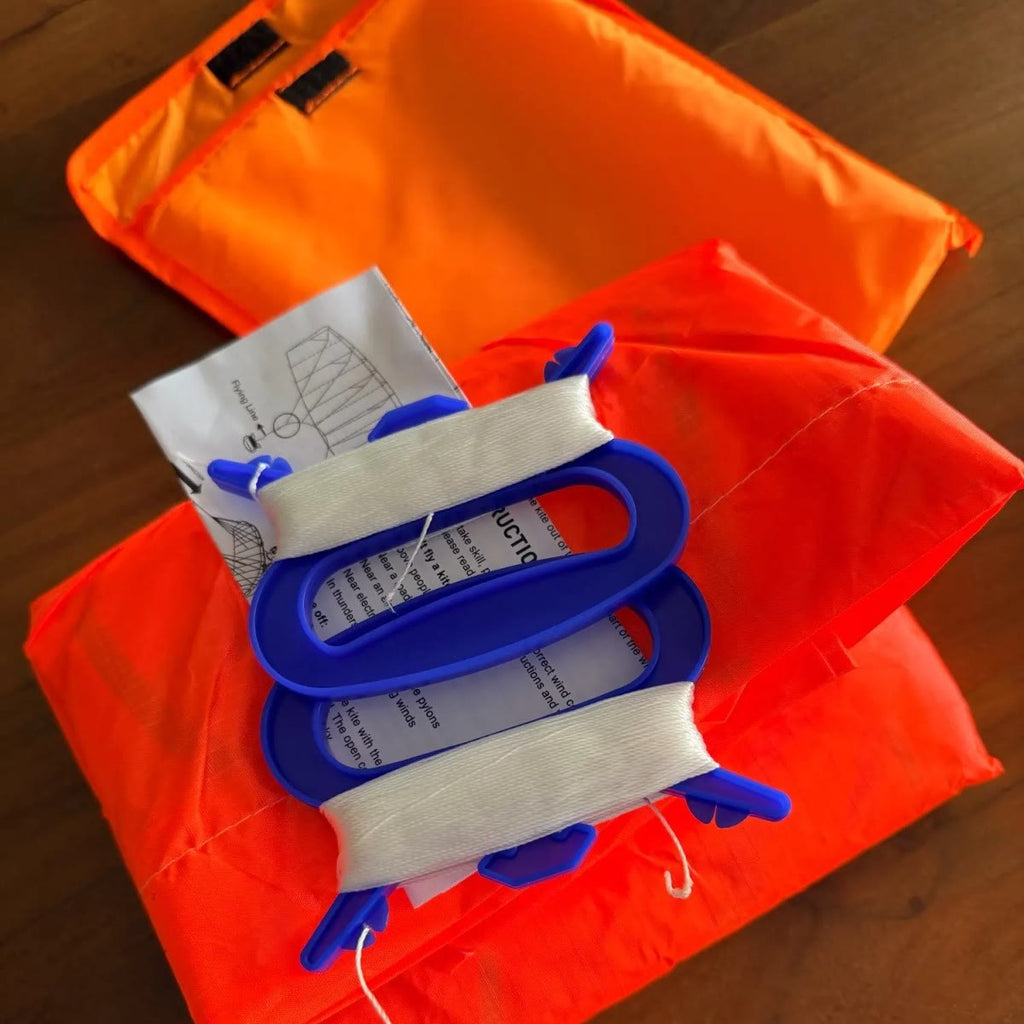 De inhoud van het pakket: oranje zakje met klittenband, opgevouwen vlieger, twee blauwe handgrepen met opgewikkelde lijn en een gebruiksaanwijzing