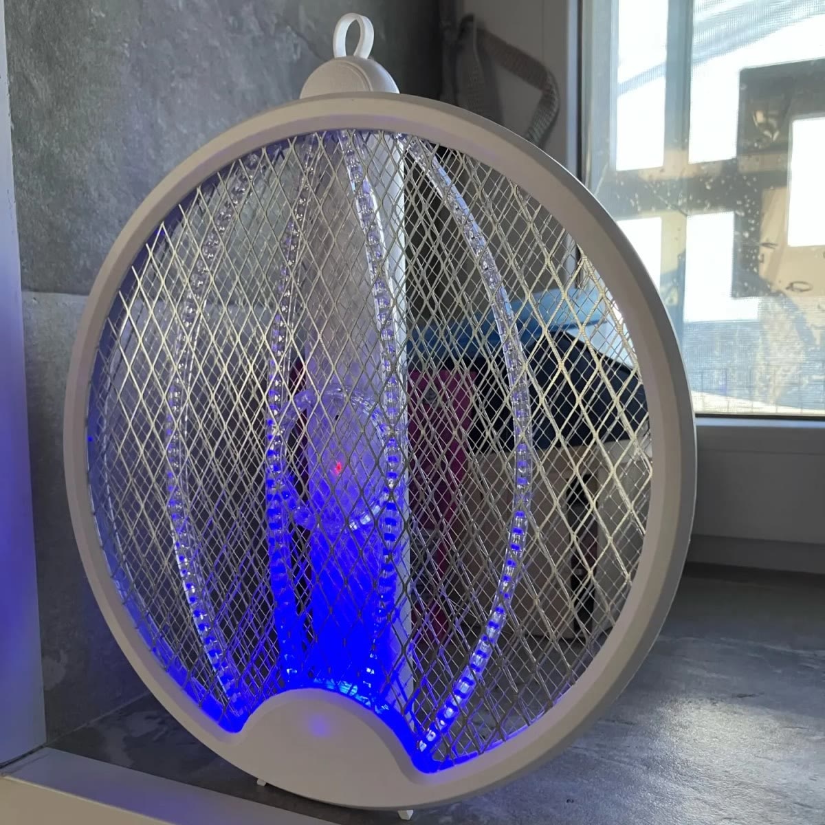 De vliegenmepper staat ingeklapt in een vensterbank, met het blauwe licht aan onder in het net