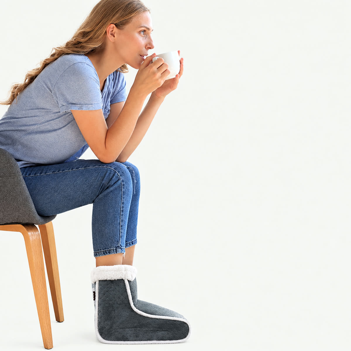 Vrouw zit op een stoel met beide voeten in de voetenwarmer en een kopje in haar hand