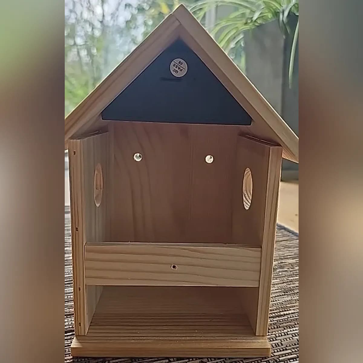 Foto van een koper: het huisje met de voorkant eraf, met de zwarte camera onder de nok, twee schroeven in de achterwand en een opening in elke zijwand