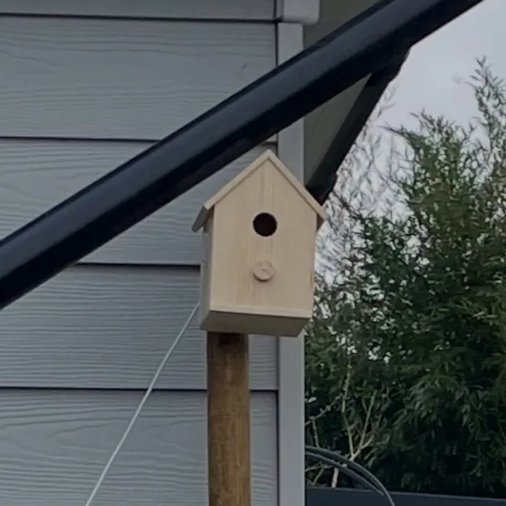 Foto van een koper: het vogelhuisje op een paal bij een huismuur, met de witte kabel die aan de achterkant naar beneden loopt