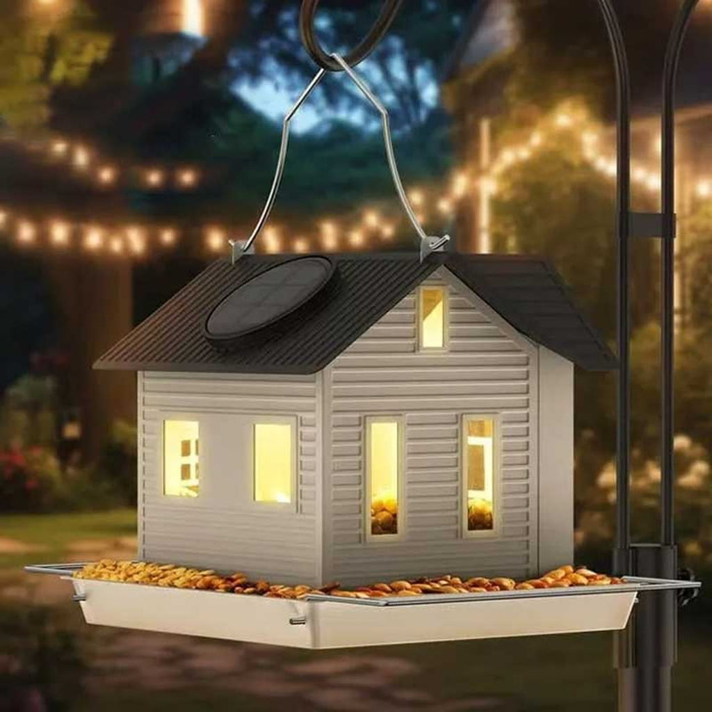 Het voederhuisje hangt in de schemering in een tuin, met licht in de raampjes en voer op het plankje