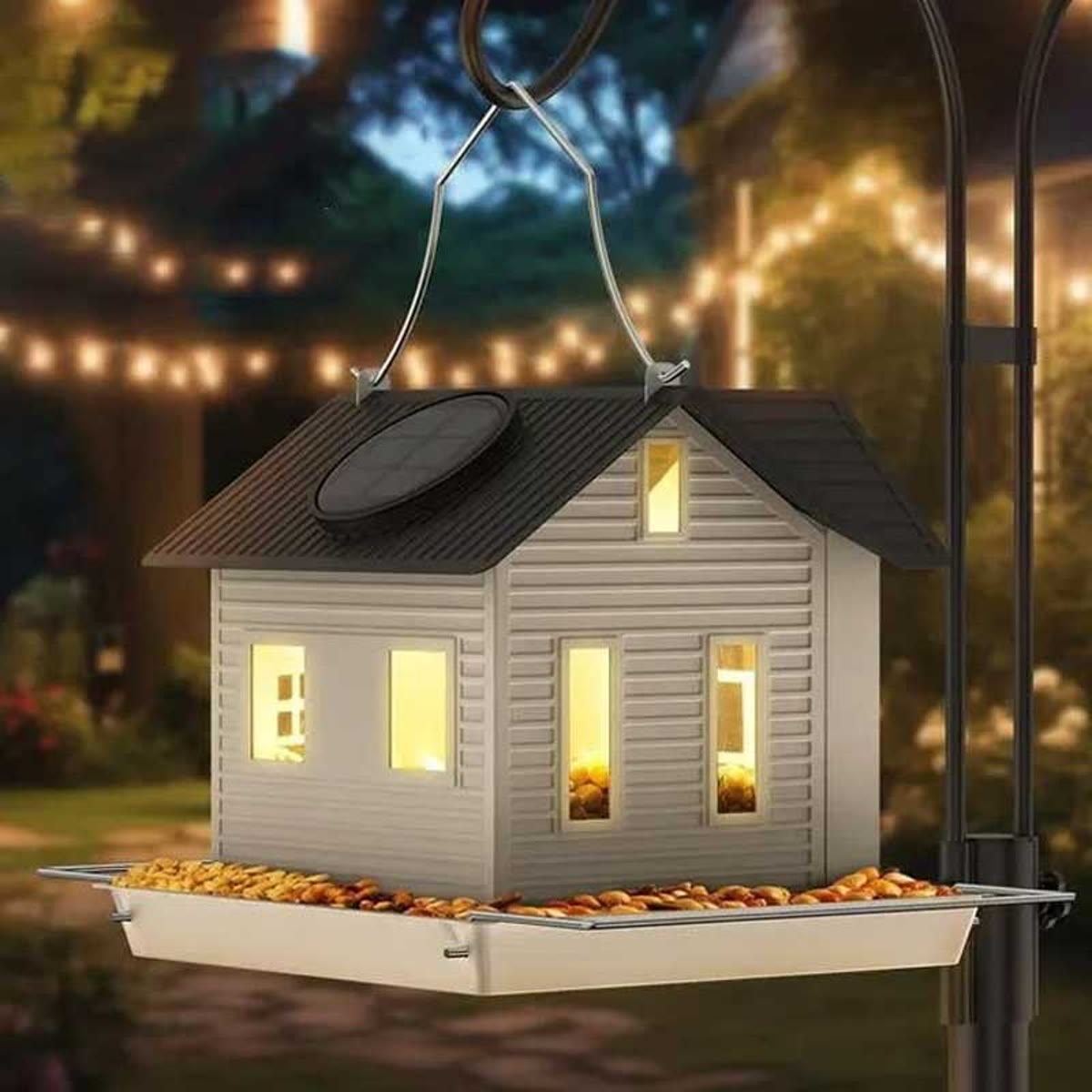 Het voederhuisje hangt in de schemering in een tuin, met licht in de raampjes en voer op het plankje