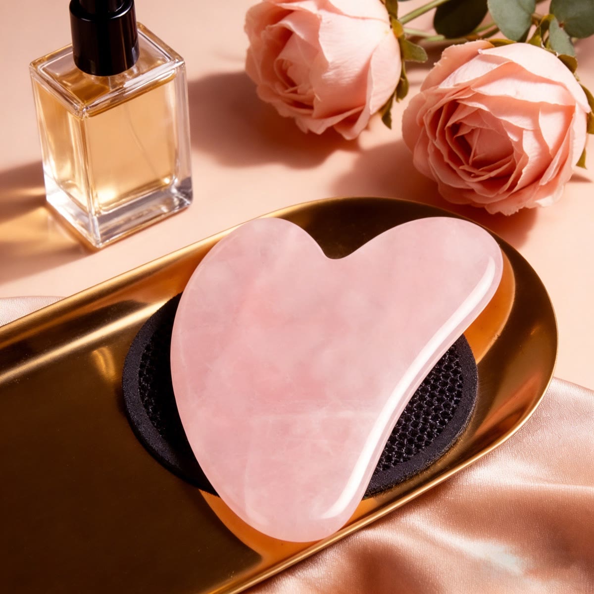 Gua sha van rozenkwarts op een gouden schaaltje naast rozen en een parfumflesje