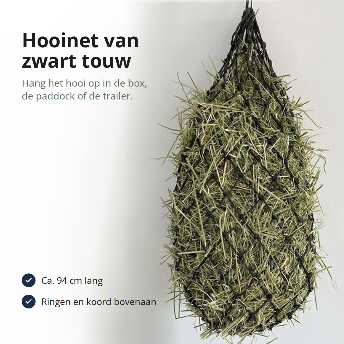 Zwart hooinet van geknoopt touw gevuld met hooi, met de tekst: hooinet van zwart touw, hang het hooi op in de box, de paddock of de trailer; ca. 94 cm lang; ringen en koord bovenaan