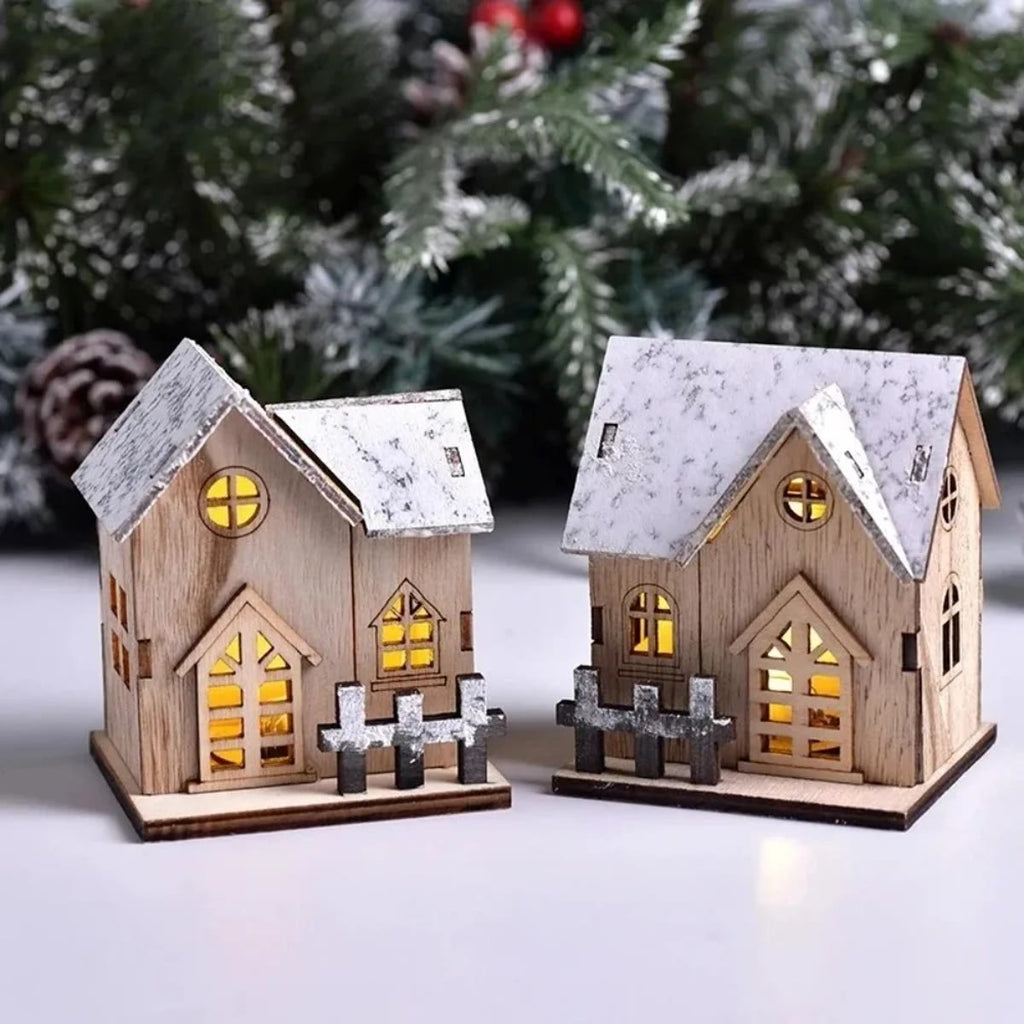 Twee houten kersthuisjes met sneeuwdak en het licht aan, op een witte tafel voor dennentakken met dennenappels