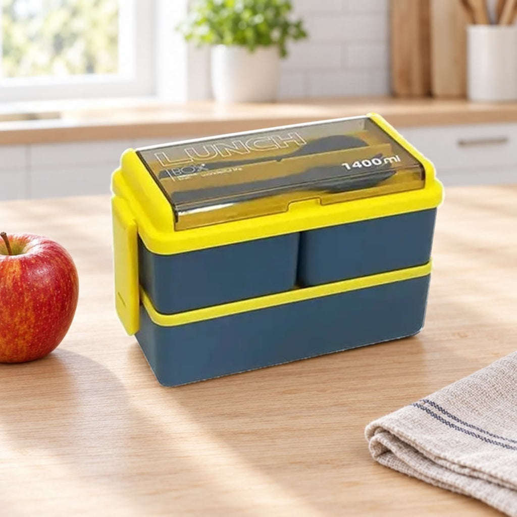 Blauwe bento box met twee lagen en geel deksel op een keukentafel naast een appel