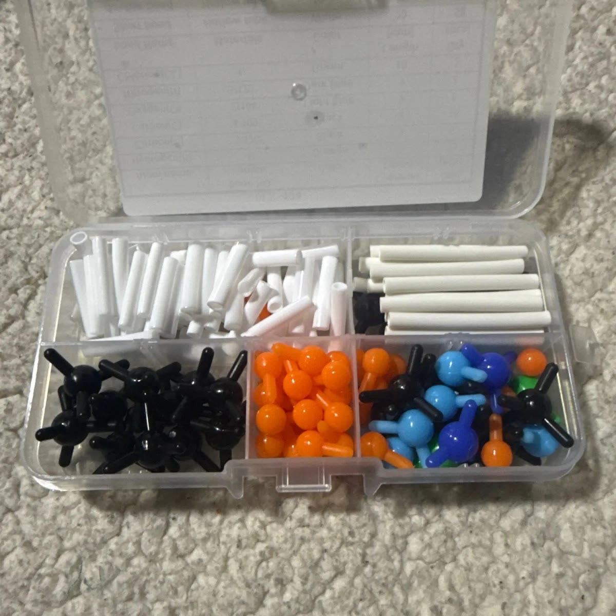 Open plastic doos met vakjes voor zwarte, oranje en blauwe atomen en korte en lange witte buisjes, gefotografeerd door een koper