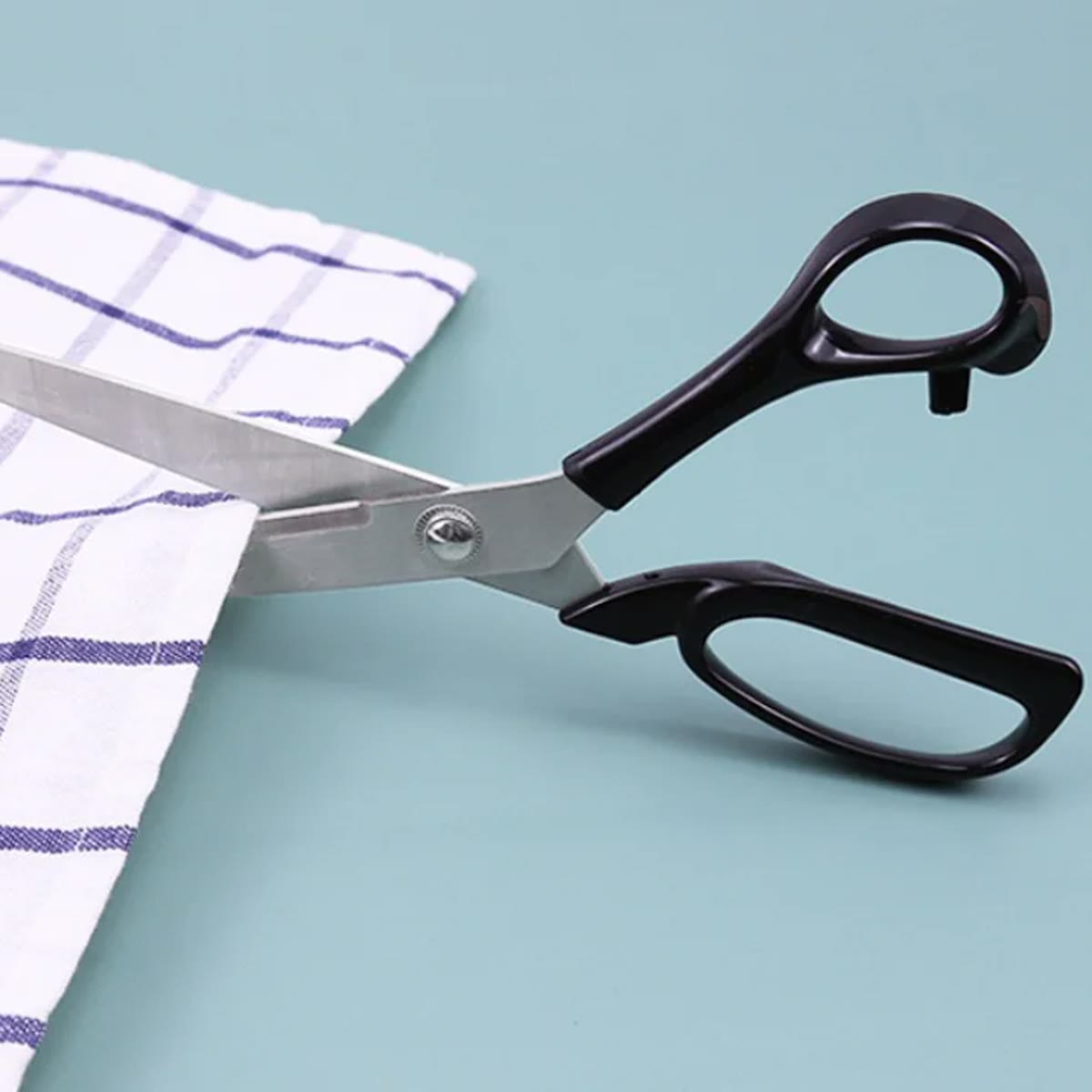 Stofschaar met zwart handvat die een geruite theedoek knipt, op een blauwgroene achtergrond