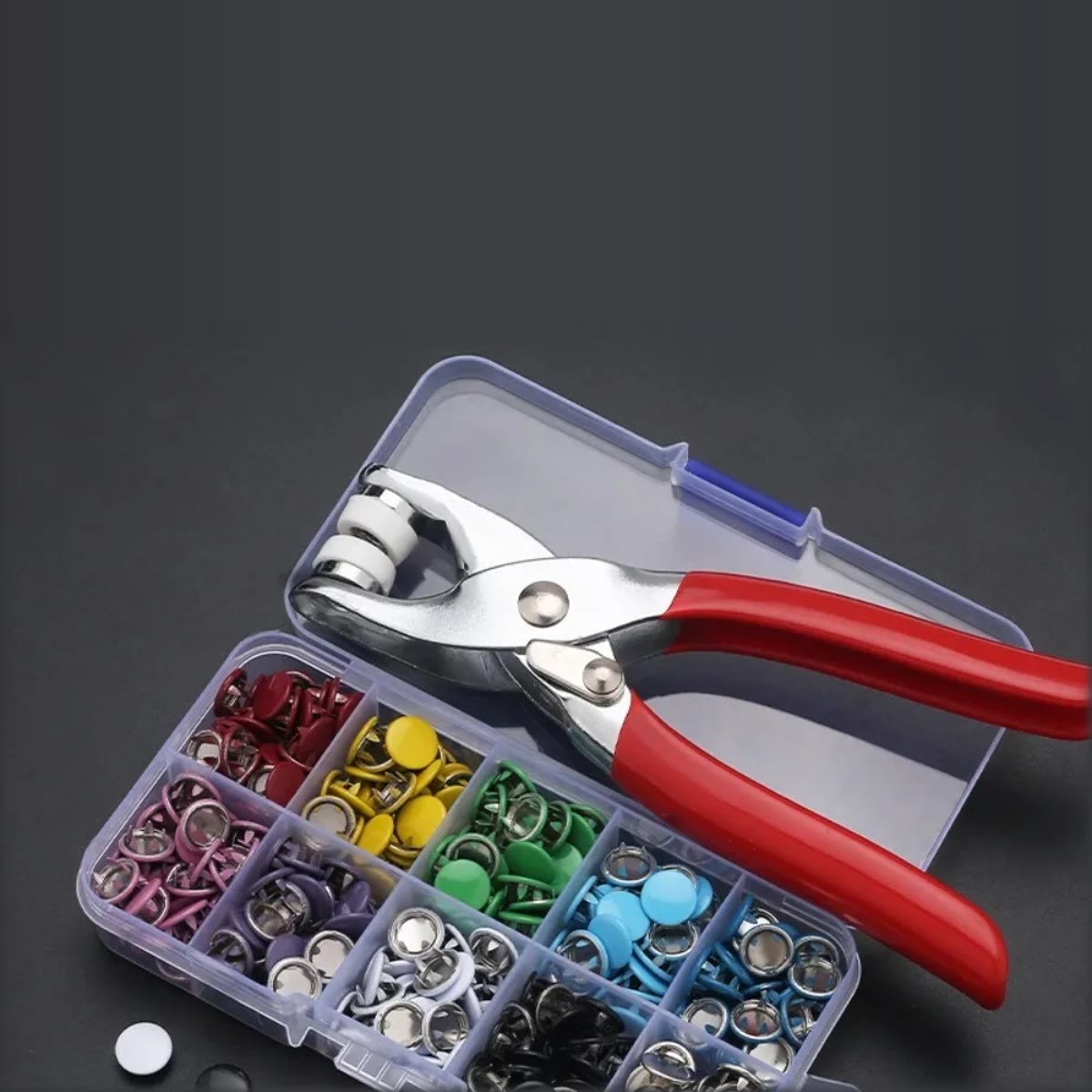 De drukknooptang met rode handvatten en de open doos met drukknopen in rood, geel, groen, blauw, roze, paars, wit en zwart