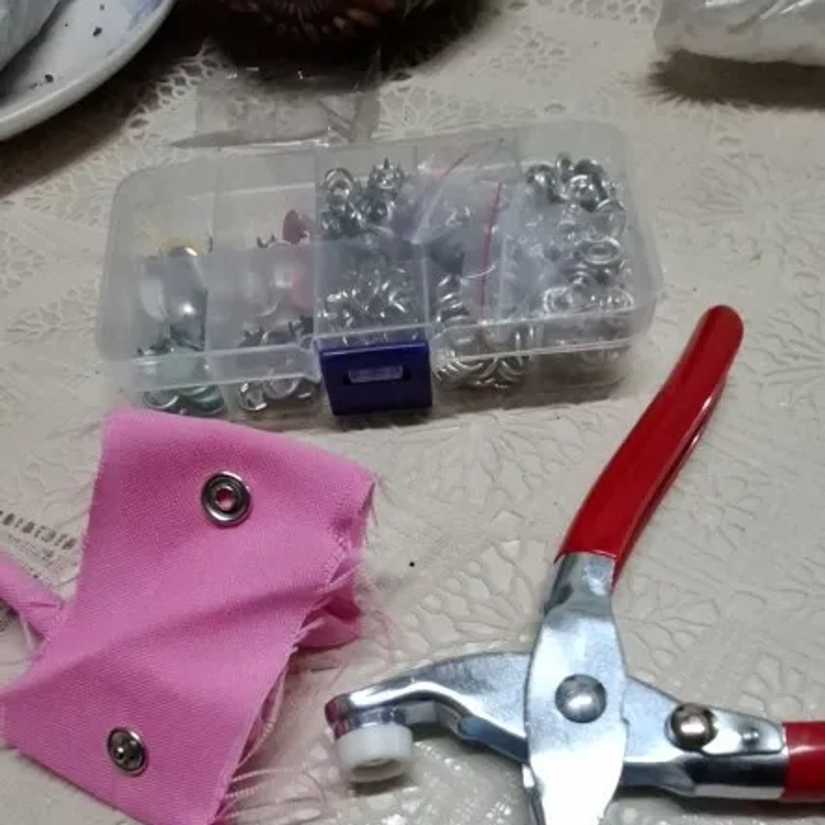 Foto van een koper: de tang, de doos en een roze lapje stof met twee vastgezette drukknopen op een tafel