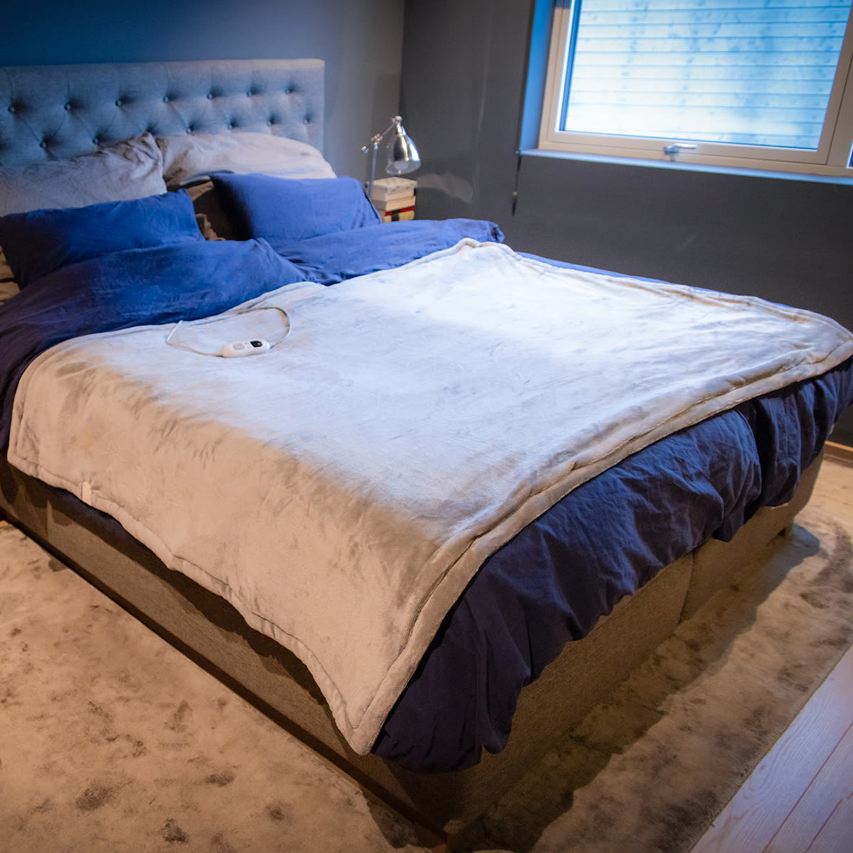 Lichtgrijze warmtedeken uitgespreid over een tweepersoonsbed met blauw beddengoed