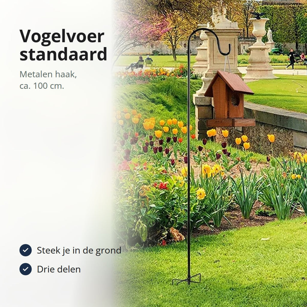 Zwarte metalen haak in een bloemenborder met een vogelhuisje aan de haak, met de tekst: vogelvoer standaard, metalen haak van ca. 100 cm, steek je in de grond, drie delen